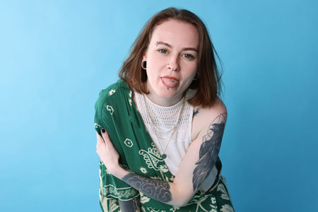 Young woman with piercings, tattoos and stylish clothes showing tongue on light blue background