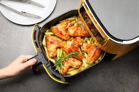 Woman opening air fryer with grilled chicken wings and French fries at gray table, top viewの写真素材