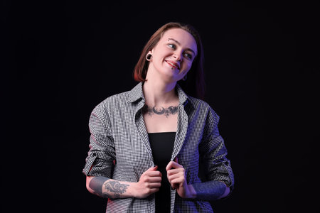 Young woman with piercings, tattoos and stylish clothes on black background
