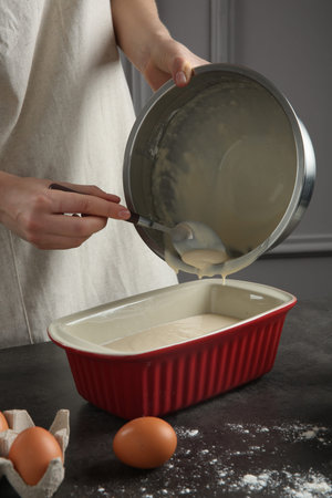 Woman pouring liquid dough from bowl into baking dish at gray table in kitchen, closeup