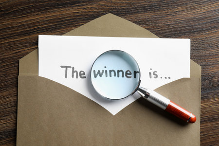 Paper note with phrase The Winner Is in envelope and magnifying glass on wooden table, top viewの写真素材