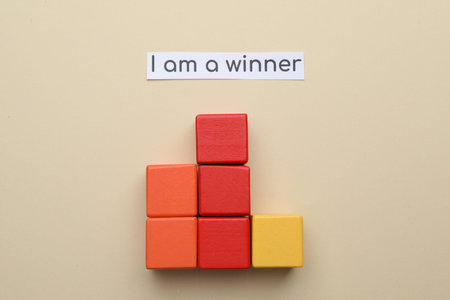 Paper note with phrase I Am A Winner and stacked cubes on beige background, flat lay