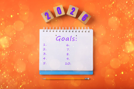New 2026 Year Goals. Inscription in notebook and cubes with numbers on orange, top view. Bokeh...