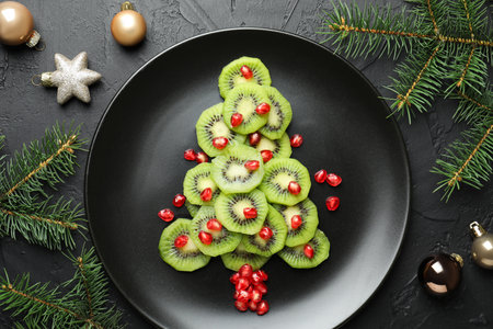 Christmas tree made of kiwi, pomegranate seeds and festive decor on black table, flat layの写真素材