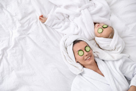 Mother and daughter in bathrobes with cucumber slices on eyes and towels wrapped around hair lying on bed, above view. Space for textの写真素材