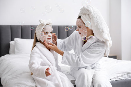 Mother and daughter in bathrobes applying face masks on bed at homeの写真素材