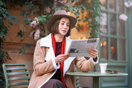 Beautiful woman in hat and coat reading newspaper at table in outdoor cafeの写真素材