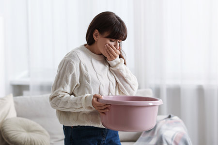 Young woman with basin suffering from nausea at homeの写真素材