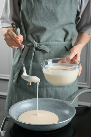 Woman pouring liquid dough from bowl into frying pan at gray table in kitchen, closeupの写真素材