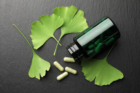 Ginkgo biloba leaves and medical bottle with pills on dark textured table, flat lay
