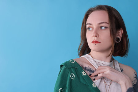 Young woman with piercings, tattoos and stylish clothes posing on light blue background, space for...