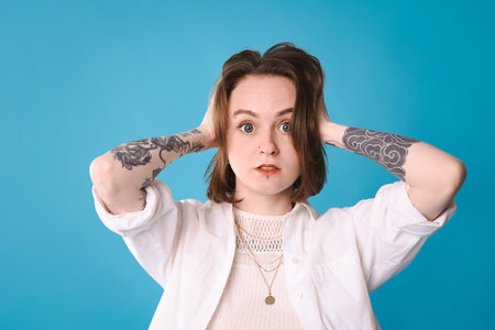 Shocked young woman with piercings, tattoos and stylish clothes posing on light blue backgroundの写真素材