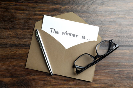 Paper note with phrase The Winner Is in envelope, pen and glasses on wooden table, flat lay