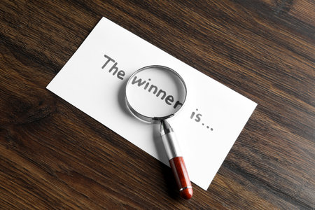 Paper note with phrase The Winner Is and magnifying glass on wooden table, above viewの写真素材