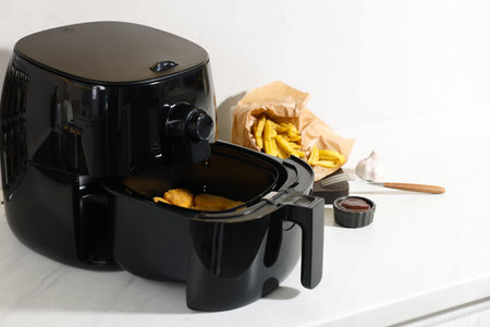 Air fryer with chicken nuggets, French fries, sauce and garlic on white marble countertop, closeup. Space for textの写真素材