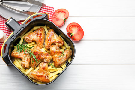 Grilled chicken wings with French fries in air fryer basket and tongs on white wooden table, flat lay. Space for textの写真素材