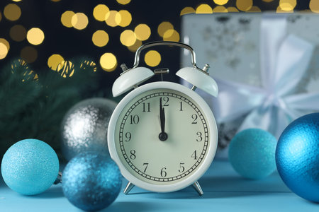 New Year celebration. Alarm clock and festive decor on light blue background against blurred lights,...