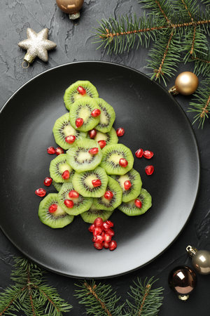 Christmas tree made of kiwi, pomegranate seeds and festive decor on black table, flat layの写真素材