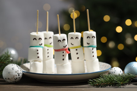 Funny snowmen made of marshmallows, sour belts and Christmas decor on wooden table against blurred lights, closeupの写真素材