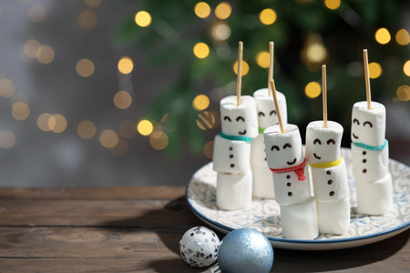Funny snowmen made of marshmallows, sour belts and baubles on wooden table against blurred lights, closeup with space for text. Christmas snacksの写真素材