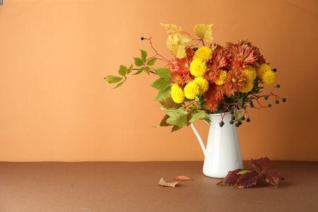 Autumn atmosphere. Vase with beautiful flowers, berries and leaves on brown table against orange background, space for textの写真素材