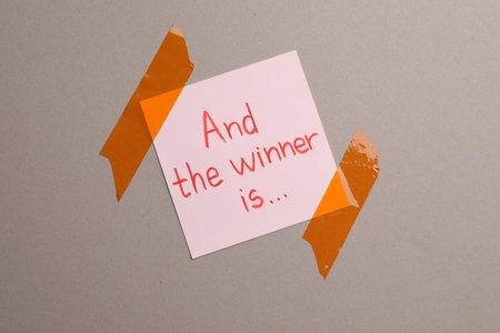 Sticky note with phrase And The Winner Is on gray background, top view
