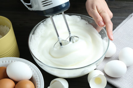 Woman whisking egg whites with hand mixer at black wooden table, closeupの写真素材