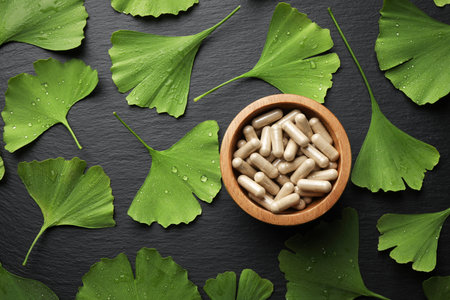 Many wet ginkgo biloba leaves and bowl of pills on dark textured table, flat layの写真素材
