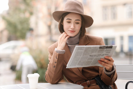 Elegant woman in hat and suit reading newspaper in outdoor cafe. Space for text