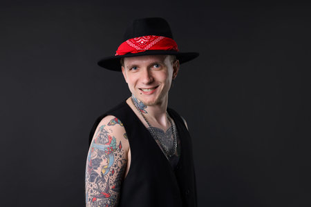 Young man with piercings, tattoos and stylish clothes posing on dark gray background