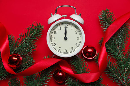 Alarm clock, fir tree branches, ribbon and baubles on red background, flat lay. New Year greeting...