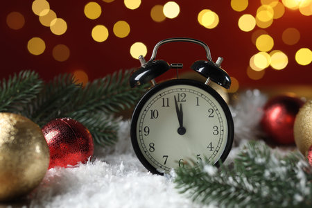 New Year celebration. Alarm clock and festive decor on artificial snow against blurred lights,...