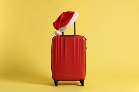 Stylish suitcase with Santa hat on yellow background. Christmas vacationの写真素材