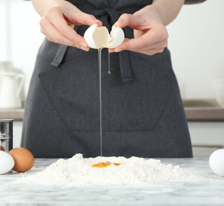 Woman cracking egg into flour at white marble table in kitchen, closeupの写真素材