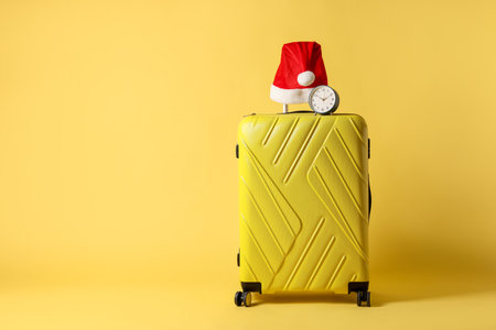 Stylish suitcase with Santa hat and alarm clock on yellow background, space for text. Christmas vacationの写真素材
