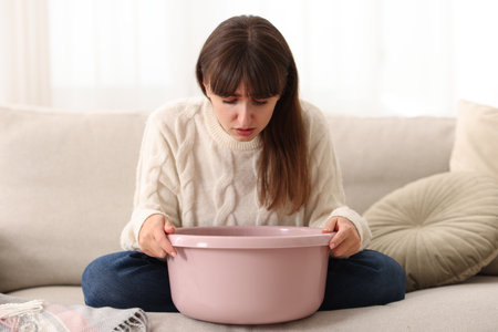 Young woman with basin suffering from nausea at homeの写真素材
