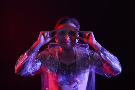 Young man with piercings, tattoos and stylish clothes posing in neon lights on black backgroundの写真素材