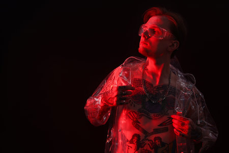 Young man with piercings, tattoos and stylish clothes posing in neon lights on black background, space for textの写真素材