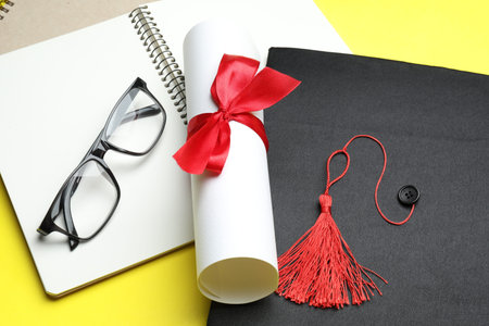 Diploma, graduation hat, glasses and notebooks on yellow background