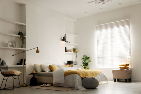 Large bed with linens, bedside tables, poufs and lamp near white wall in bedroomの写真素材