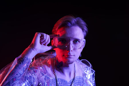 Young man with piercings, tattoos and stylish clothes posing in neon lights on black background