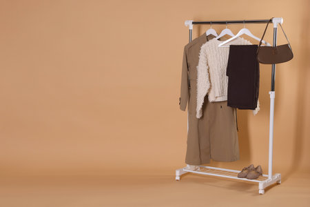 Different stylish clothes hanging on rack, bag and shoes against beige background, space for textの写真素材