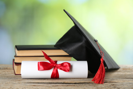 Diploma, graduation hat and stack of books on wooden table against blurred background, closeupの写真素材