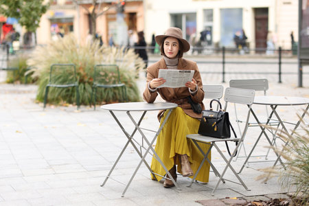 Elegant woman in hat, skirt and suit reading newspaper in outdoor cafe. Space for textの写真素材
