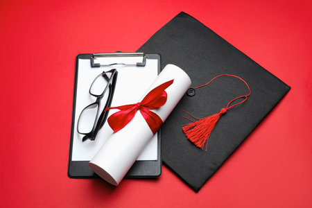 Diploma, graduation hat, glasses and clipboard on red background, above viewの写真素材