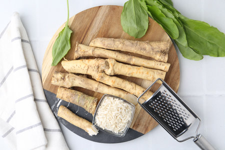 Whole and grated horseradish roots, grater and green leaves on white tiled table, top viewの写真素材