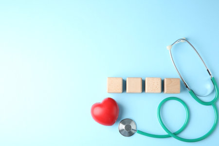 Stethoscope, heart figure and blank wooden cubes on light blue background, flat lay. Mockup for designの写真素材