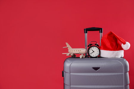 Christmas vacation. Gray suitcase with Santa hat, alarm clock and wooden plane on red background, closeup. Space for textの写真素材