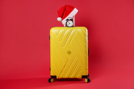 Christmas vacation. Yellow suitcase with Santa hat and alarm clock on red backgroundの写真素材
