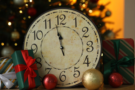 Wall clock, baubles and gift boxes on table against blurred lights, closeup. New Year greeting card